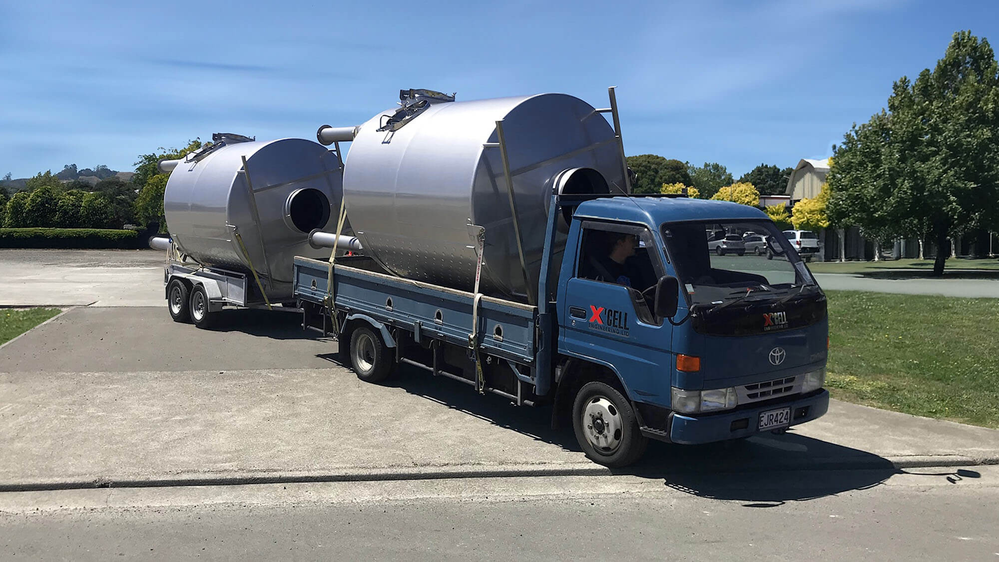 about-2 A blue X'Cell Engineering flatbed truck and trailer transports two large, horizontal stainless-steel wine tanks down a paved road on a sunny day.