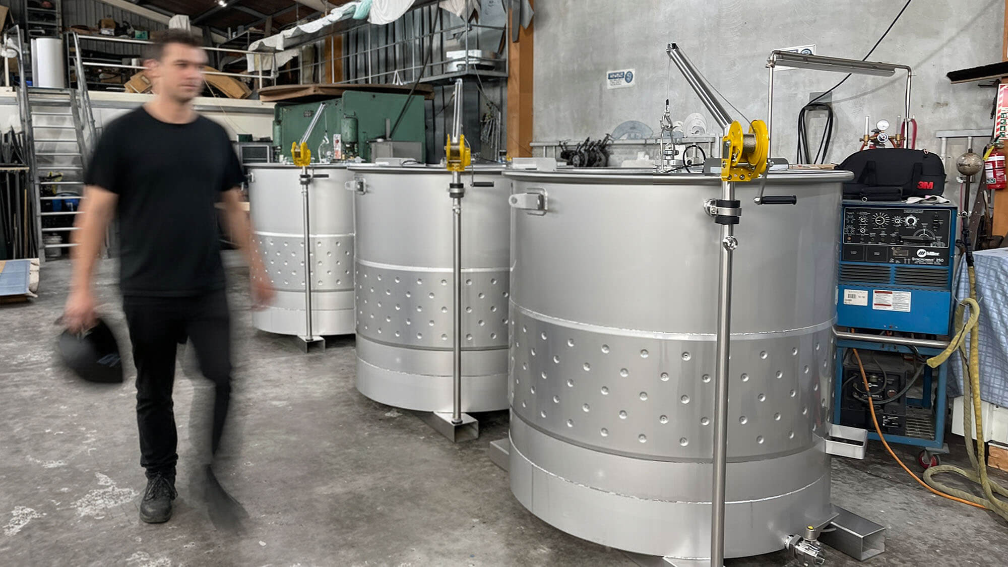 about-3 Three new stainless-steel wine tanks with dimple jackets and davit arms stand in a workshop as a worker walks past.