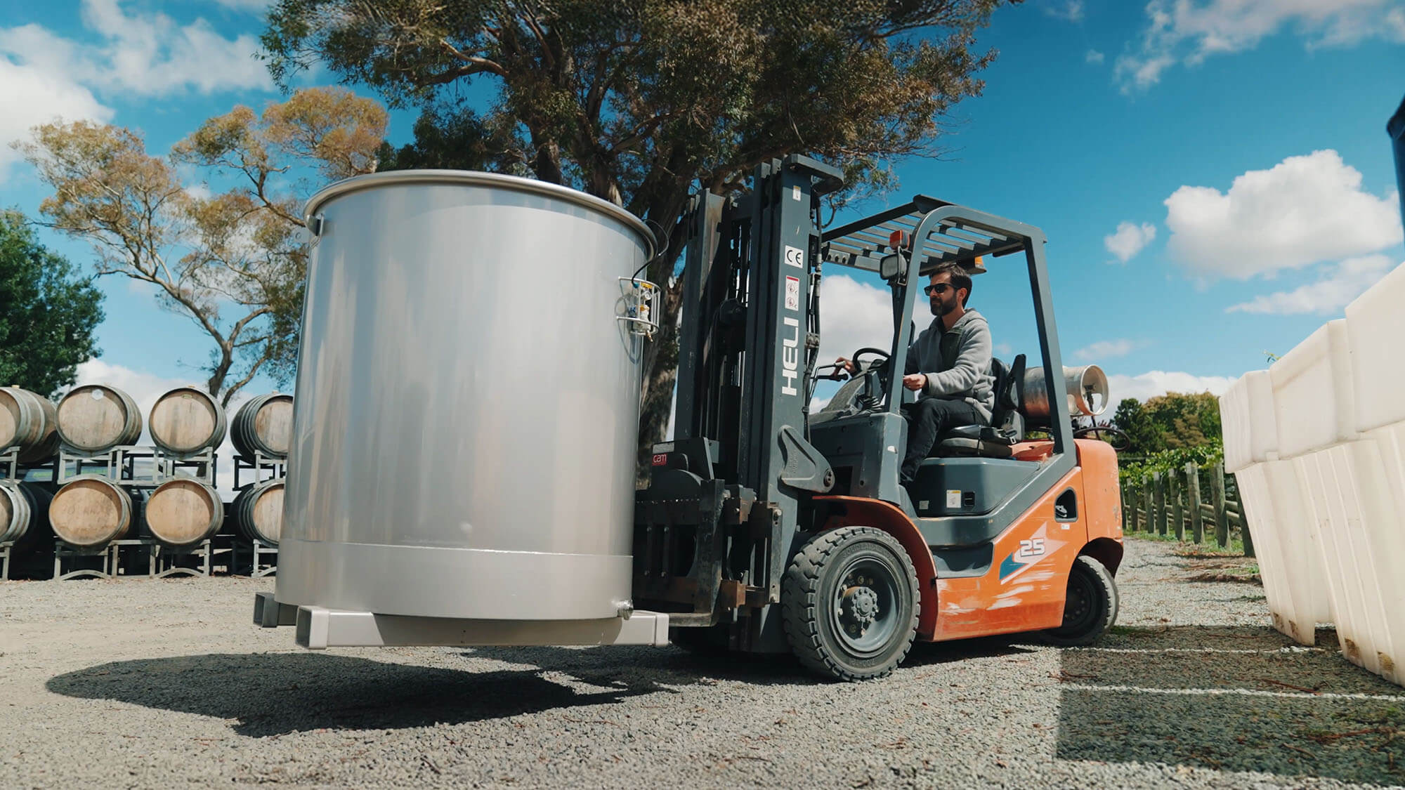 multi-08 A man drives an orange forklift, carrying a large X'Cell Multi stainless steel wine tank, across a gravel lot past stacked wine barrels under a blue sky.
