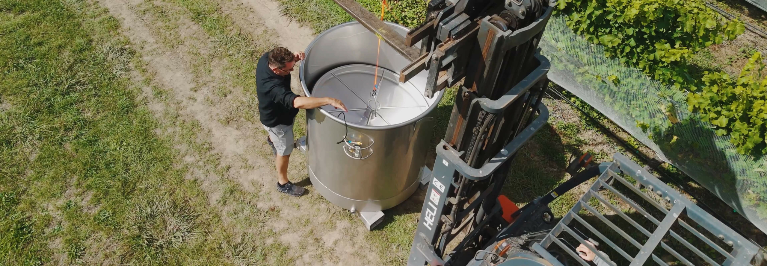 multi-hero An overhead shot of a man adjusting a stainless steel tank as a forklift lowers it into place in a vineyard.