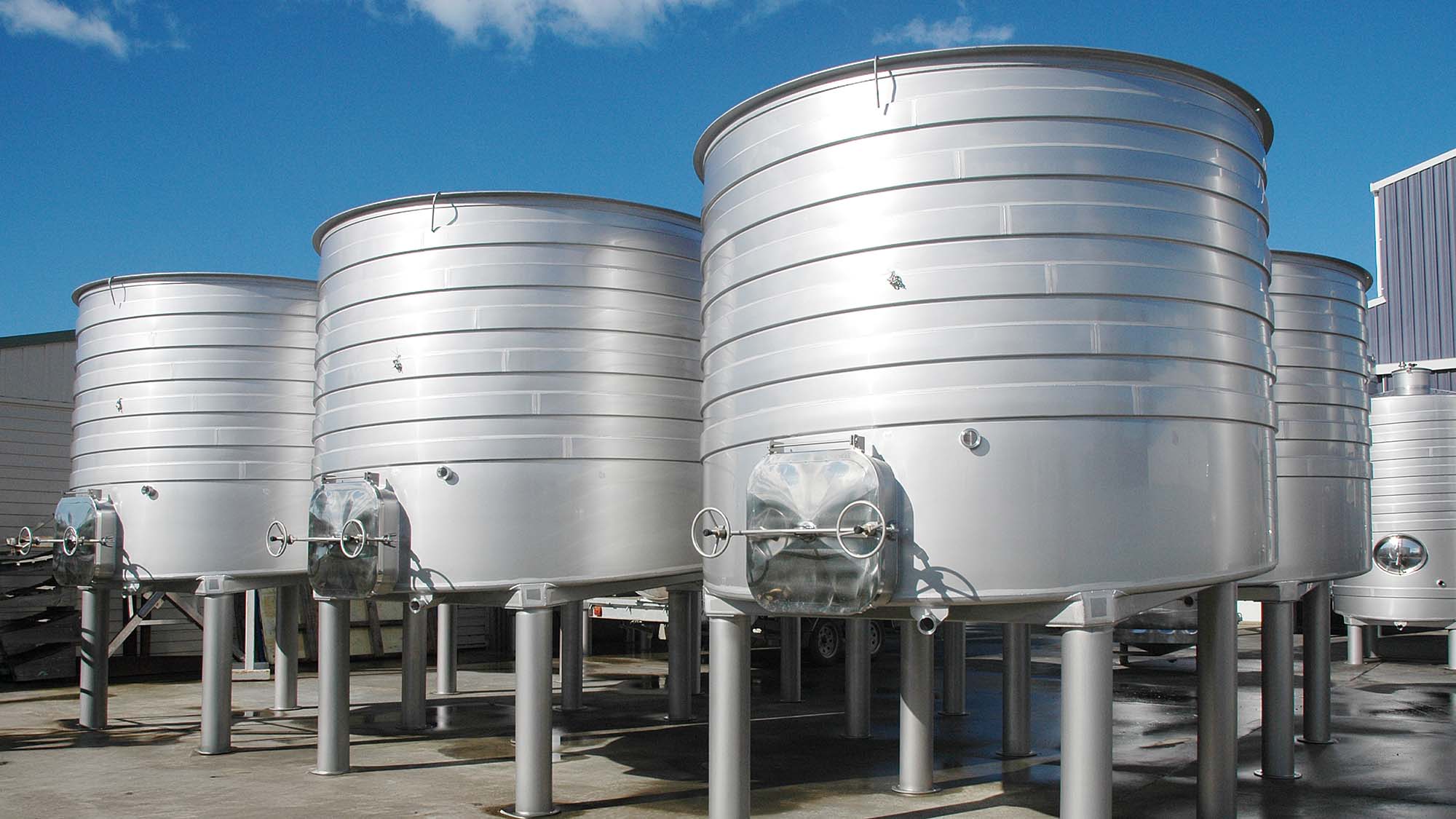 about-4 A row of large stainless steel tanks positioned outdoors, featuring elevated support legs and rectangular manway doors.