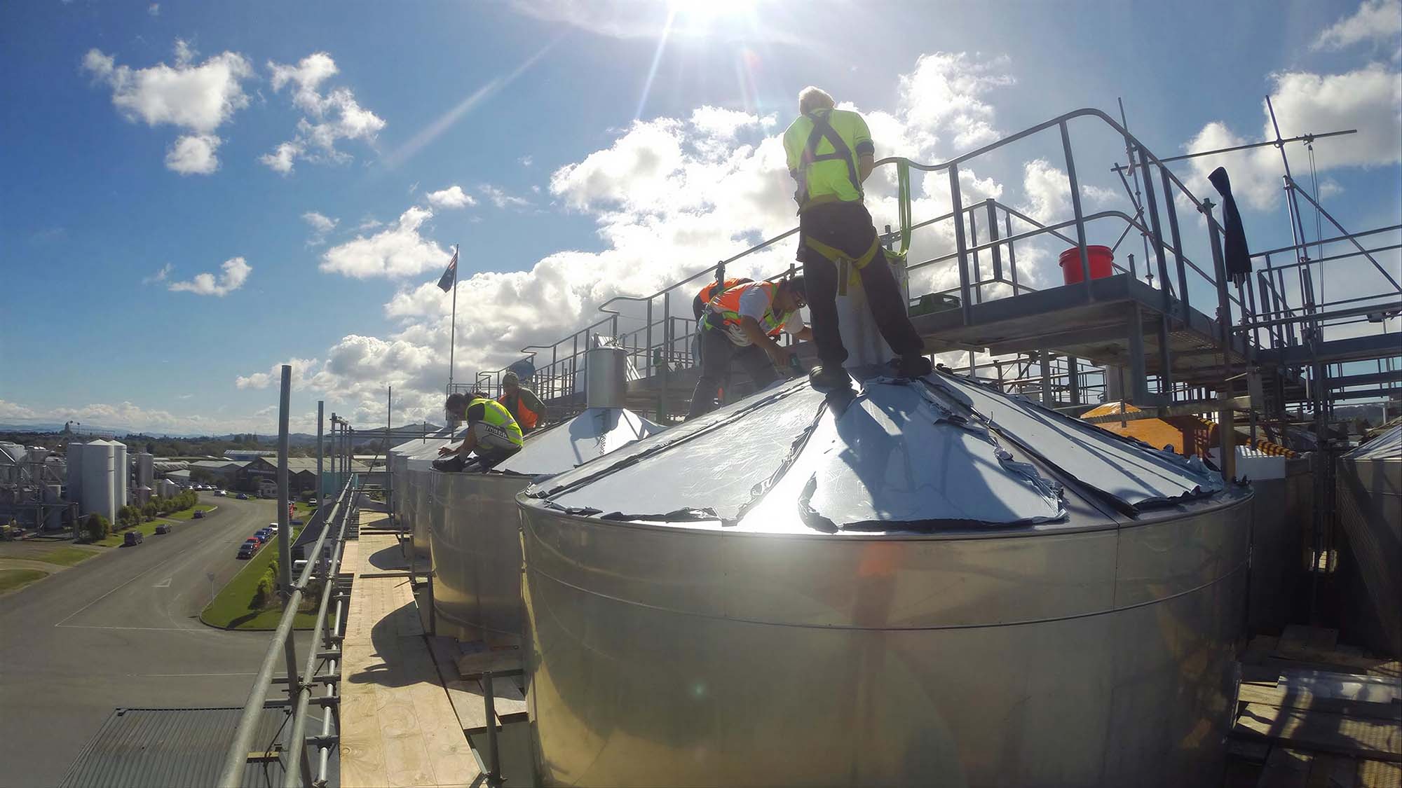 about-5 Workers in safety harnesses atop large stainless steel tanks, installing the conical roofs from a scaffolding walkway under a bright sky.